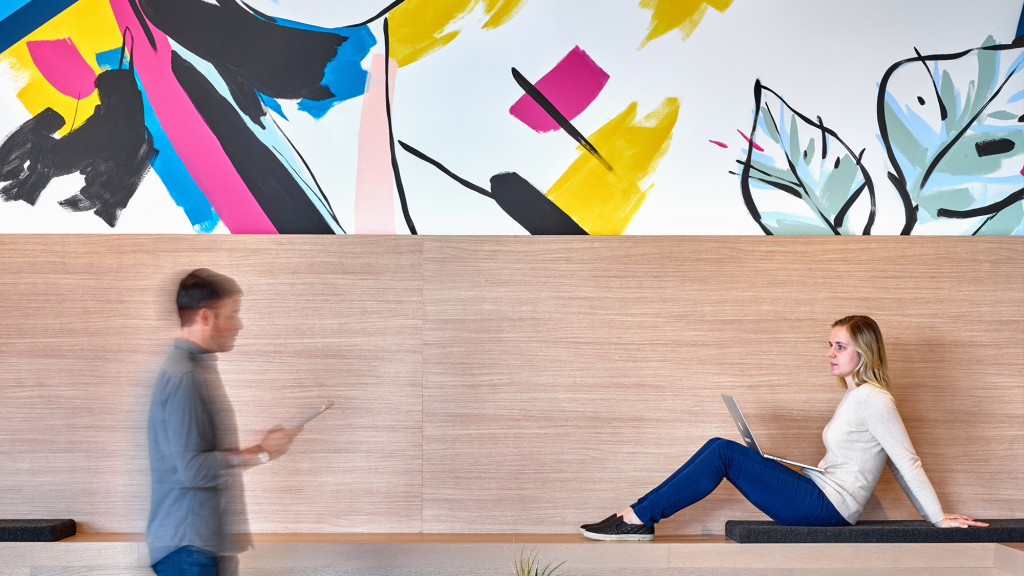 A man and woman sitting on a bench with a wall with art.